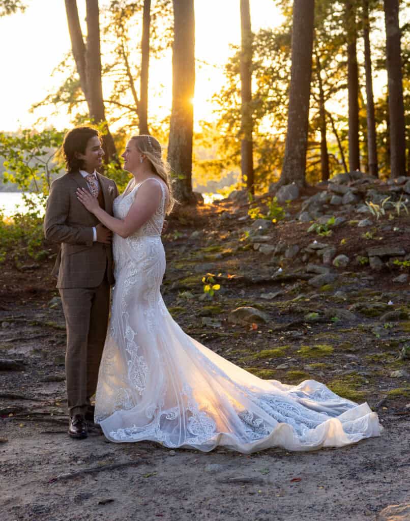 Bride in lace gown and groom in brown suit stand in wooded area at sunset, warm light filtering through trees.