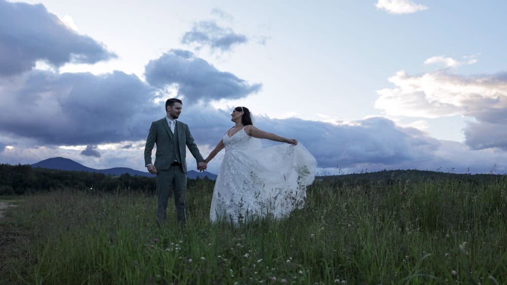 Questions to Ask a Wedding Photographer A bride and groom stand in a grassy field holding hands, with the bride lifting her dress and a cloudy sky in the background.